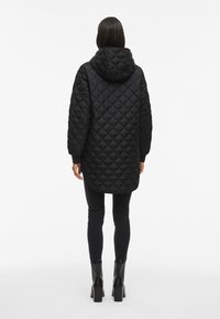 Abrigo largo acolchado negro con capucha, con un patrón de diamantes, puños acanalados y un ajuste relajado, combinado con jeans skinny negros y botas.