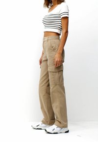 Femme portant un pantalon cargo beige, un t-shirt court blanc à rayures, et des baskets blanches, se tenant devant un fond blanc uni.