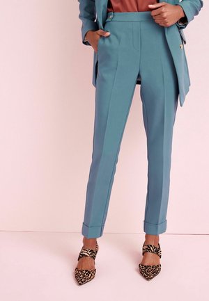 Kvinde iført turkis skræddersyede bukser med opsmøg, matchende blazer og leopardmønstrede spidse hæle mod en lyserød baggrund.