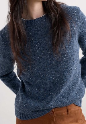 Femme portant un pull tricoté bleu tacheté et un pantalon marron, avec de longs cheveux foncés couvrant partiellement le pull.
