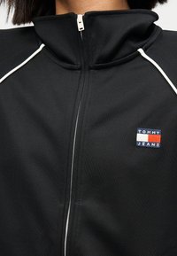 Μαύρο μπουφάν με φερμουάρ από λείο ύφασμα. Διαθέτει ψηλό γιακά, λευκές λεπτομέρειες και μικρό κεντητό λογότυπο σε μπλε, κόκκινο και λευκό.