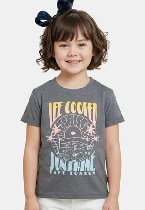 Jeune enfant souriant aux cheveux courts et bouclés avec un nœud bleu, portant un t-shirt gris avec un motif graphique pastel "Lee Cooper Sunshine East London".