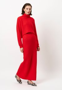 Rode gebreide coltrui gecombineerd met een bijpassende lange rok. De stof heeft een zachte textuur. Het model draagt zwart-wit gemusterde schoenen.