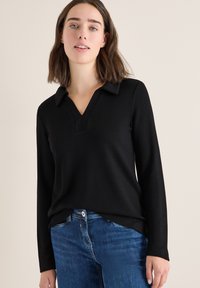 Mujer con cabello castaño de longitud media, vistiendo una blusa negra de manga larga con cuello y jeans azules, de pie frente a una pared clara y sencilla.