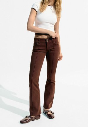 Jeans évasés marron avec des coutures contrastées, taille haute, et deux poches avant. Assortis à une chemise blanche courte et des chaussures plates.