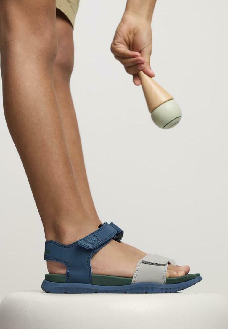 Sandalias ajustables en azul y gris con suela de goma, que cuentan con una tira más ancha y acentos texturizados, colocadas sobre una superficie blanca.