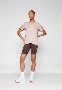 Kvinne står med hendene på hoftene iført en lys mauve t-skjorte med korte ermer, mørkebrun sykkelshorts, hvite sokker og hvite løpesko.