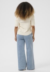 Žena s kudrnatými vlasy, oblečená v krémovém pleteném topu s krátkým rukávem a světle modrých širokých džínách, stojící čelem k objektivu na jednobarevném pozadí.