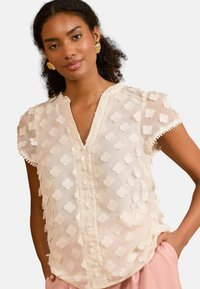 Helle beige Bluse mit strukturierten floralem Applikationen, V-Ausschnitt, kurzen Ärmeln und dekorativem Saum. Weicher, transparenter Stoff mit Knopfverschluss.
