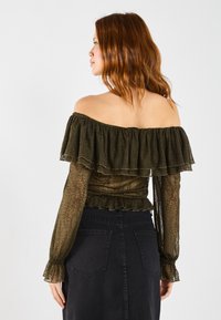 Off-shoulder topp i mørkegrønn med volanger, teksturert stoff, gjennomsiktige lange ermer og tettsittende design, kombinert med et svart skjørt.