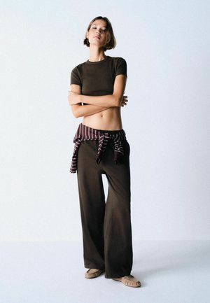 Junge Frau steht mit verschränkten Armen, trägt ein braunes Crop-Top, weite Hosen, ein gestreiftes Hemd um die Taille gebunden und beige flache Schuhe.