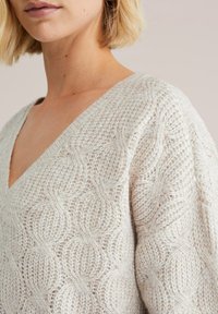 Maglione lavorato a maglia di colore beige chiaro con scollatura a V e motivo testurizzato di cerchi in rilievo. Materiale morbido e accogliente con una vestibilità ampia e finitura testurizzata.