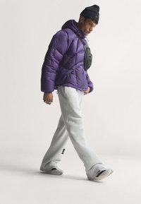 Mann trägt eine lila Steppjacke, schwarze Mütze, weiße Sneakers, graue Jogginghose und eine schwarze Umhängetasche und geht vor einem einfarbigen Hintergrund.