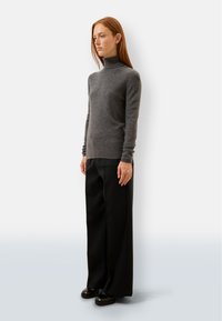 Grauer Wollrollkragenpullover mit langen Ärmeln, kombiniert mit schwarzen weit geschnittenen Hosen und schwarzen Schuhen, vor einem neutralen Hintergrund.