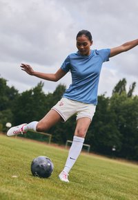Gaiši zila sporta krekliņa un baltu šortu ar krāsainām svītrām. Spēlētājs driblē melnu futbolu uz zāliena. Baltas zeķes un rozā akcentiem aprīkotas futbolu kurpes.