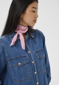 Camicia in denim blu con bottoni dorati e due tasche sul petto, abbinata a una sciarpa strutturata rosa chiaro annodata attorno al collo.