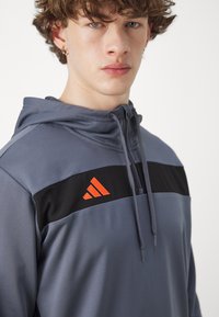 Grauer Hoodie mit schwarzem Panel und orangefarbener Adidas-Logo. Verfügt über eine verstellbare Kapuze, lange Ärmel und glattes, leichtes Material.