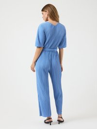 Kvinne iført en belted, kortermet, blå plisserte jumpsuit med brede ben, sett fra baksiden, stående på en hvit bakgrunn.