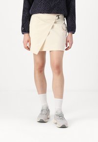Beige omslagskjol gjord av lättviktsmaterial, med en sidoficka och bältesöggla. Bärs med grå sneakers och vita strumpor.