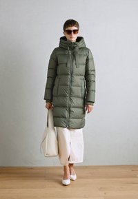 Olijfgroene pufferjas met een capuchon, ritssluiting en elastische manchet, gedragen over witte wijde broek, gecombineerd met witte schoenen en een schoudertas.