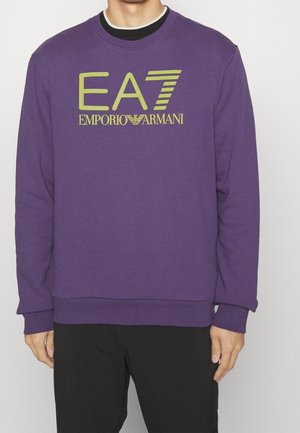 Henkilö, jolla on päällään violetti collegepaita, jossa on vihreä "EA7 Emporio Armani" -logo rinnassa, sekä mustat housut, seisomassa yksinkertaisen valkoisen taustan edessä.