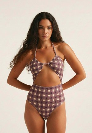 Mujer con un bañador halter morado con estampado de estrellas y recortes, de pie con las manos detrás de la espalda sobre un fondo claro y liso.