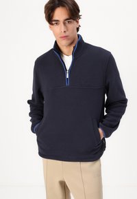 Tengeri kék fleece pulóver negyed cipzárral, kontrasztos kék gallérral, oldalsó zsebekkel és diszkrét logóval a mellkason.