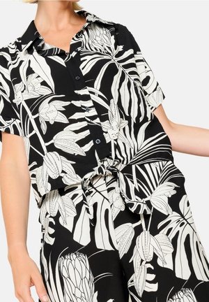 Kobieta ubrana w czarno-białą koszulę w kwiatowy wzór z krótkimi rękawami oraz dopasowane spodnie, związane w pasie węzłem.