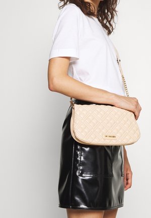 Kvinde iført hvid t-shirt og sort lædernederdel holder beige quiltet crossbody-taske med guldkæde og logoplaket.