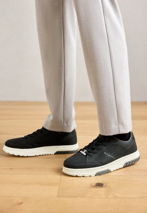 Benen in lichtbeige broek en zwarte Bugatti sneakers met witte zolen staan op een houten vloer.