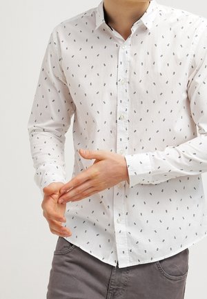 Muž v bílé košili na knoflíky s malým černým potiskem a šedých kalhotách si pravou rukou upravuje manžetu na levém rukávu.