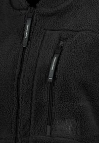 Schwarze Fleecejacke mit strukturierter Außenseite, zwei Reißverschlusstaschen und einem gestickten Logo. Die Reißverschlussanhänger sind mit "SUPREM CLTHNG" beschriftet.