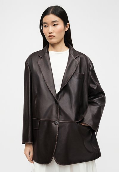 Νεαρή γυναίκα με ίσια μαύρα μαλλιά φοράει υπερμεγέθη καφέ δερμάτινο σακάκι πάνω από λευκό τοπ, κοιτάζοντας ελαφρώς προς το πλάι με φόντο απλό.