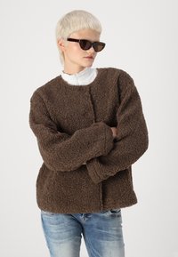 Veste marron texturée avec un design en peluche, col rond et deux grands boutons. Portée sur une chemise blanche à col haut et un jean bleu.
