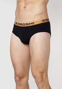 Schwarze Unterhosen aus weichem Stoff mit einem goldenen elastischen Bund, auf dem der Text "KOROSHI" aufgedruckt ist. Entworfen für eine enge Passform.
