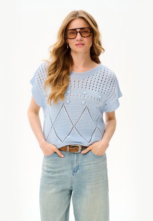 Kobieta o długich falowanych włosach, nosząca oversize brązowe okulary przeciwsłoneczne, jasnoniebieski sweterek dzianinowy z wzorami diamentów, wpuszczony do wysokich jasnych jeansów.