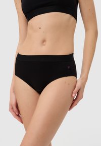 Frau trägt schwarze hochtaillierte Unterwäsche und einen passenden schwarzen Sport-BH, steht vor einem einfarbigen hellen Hintergrund.