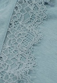 Světle modrá látka s jemným krajkovým lemem a složitým květinovým vzorem podél jednoho okraje. Měkká textura s jemnými detaily třepení.