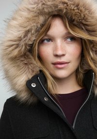 Mladá žena s pehami, oblečená v tmavom kabáte s veľkou huňatou kapucňou, pozerajúca sa mierne nabok.