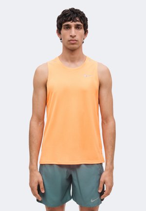 Jonge man staat recht naar voren en draagt een oranje mouwloos sportshirt en zeegroene korte broek met een klein Nike-logo, neutrale achtergrond.