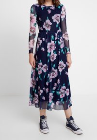 Robe bleue marine à motifs floraux avec manches longues en tulle transparent et jupe fluide. Elle présente des motifs floraux roses et verts. Associée à des baskets montantes bleu marine.