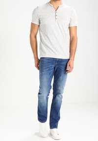 Maglietta henley grigio chiaro a maniche corte, con placket a tre bottoni, abbinata a jeans in denim blu e sneaker bianche.