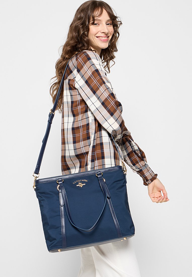 Sac tote en nylon bleu marine avec doubles poignées et bandoulière amovible. Présente un logo doré et un design minimaliste. Texture lisse.