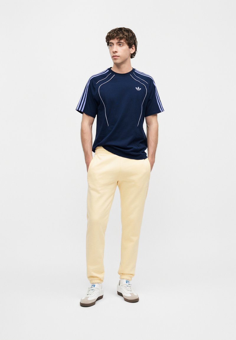 Young man standing with hands in pockets wearing a navy blue Adidas T-shirt, beige sweatpants, and white sneakers on a plain background.