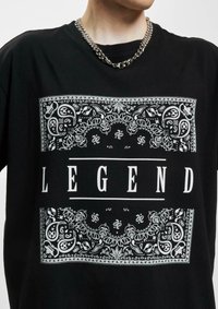 Juodas medvilninis marškinėliai su baltu grafikiniu spausdinimu, papuoštu įmantriais raštais ir žodžiu "LEGEND" ryškiomis raidėmis centre.