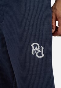 Marineblaue, gerippte Jogginghosen mit einem gestickten Logo in Weiß auf dem Oberschenkel. Das Design hebt die strukturierte Oberfläche und die lässige Passform hervor.