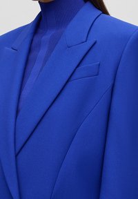 Niebieska marynarka o dopasowanym kroju. Gładka tkanina, ostre klapy i kieszeń na piersi. Warstwowy prążkowany golf w tym samym odcieniu niebieskiego pod spodem.