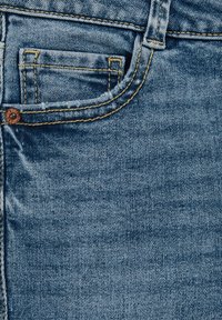 Nahaufnahme von Denim-Stoff in einem hellblauen Waschung mit subtilen Verblassungen, sichtbaren gelben Nähten und einem kleinen Detail der vorderen Tasche.