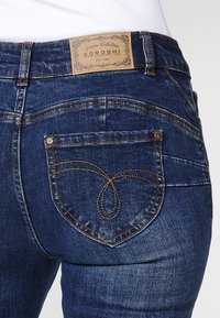 Temno modri kavbojke z edinstvenim žepom v obliki srca na hrbtni strani, z zlatim šivanjem in blagovno znamko na usnjenem patu v pasu.