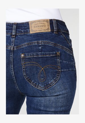 Ciemnoniebieskie dżinsy z odznaczającą się kieszenią w kształcie serca z tyłu, ozdobione złotym przeszyciem i skórzaną naszywką marki w pasie.